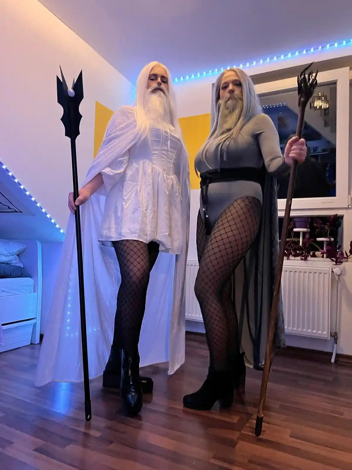 Two women in a room, both wearing long white wigs and fake beards, holding staffs. One wears a white robe and fishnet…