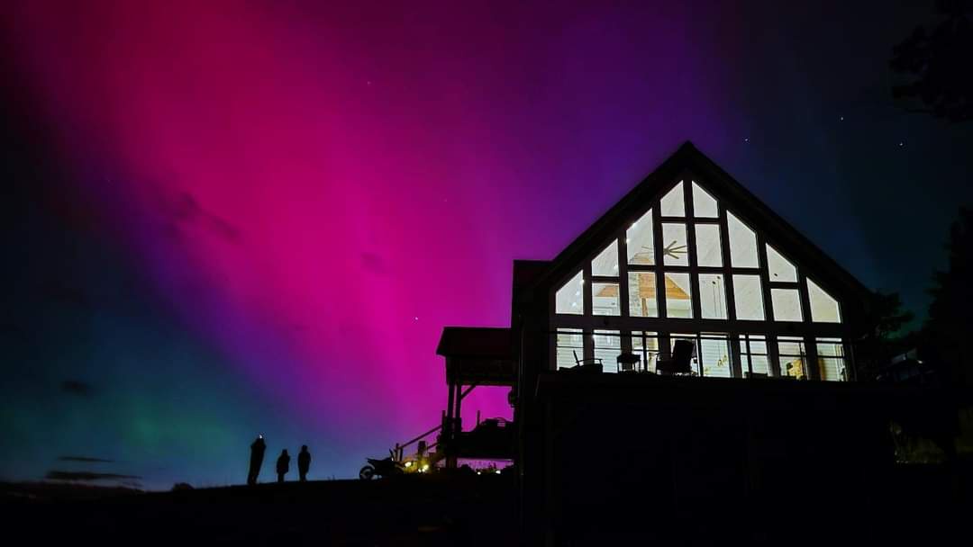 A nighttime photo shows a large, modern house with a glass-paned gable roof, its interior warmly lit, silhouetted against a…