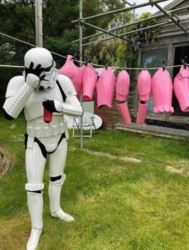 A person in a full white stormtrooper costume stands in a grassy backyard, holding a red can and covering their face with…