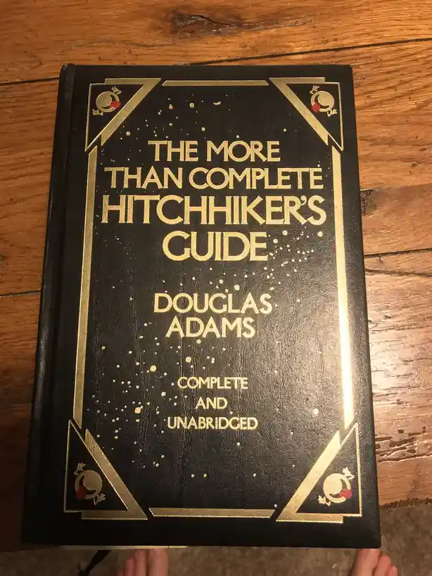 A black book with gold lettering and decorative borders sits on a wooden floor. The cover reads 'THE MORE THAN COMPLETE…