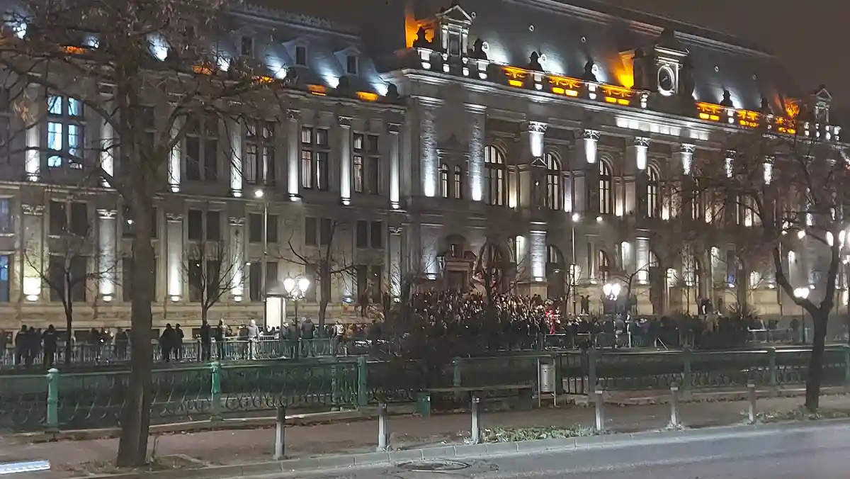 A nighttime photo of a large, ornate building with classical architecture, illuminated by spotlights, with a crowd gathered…