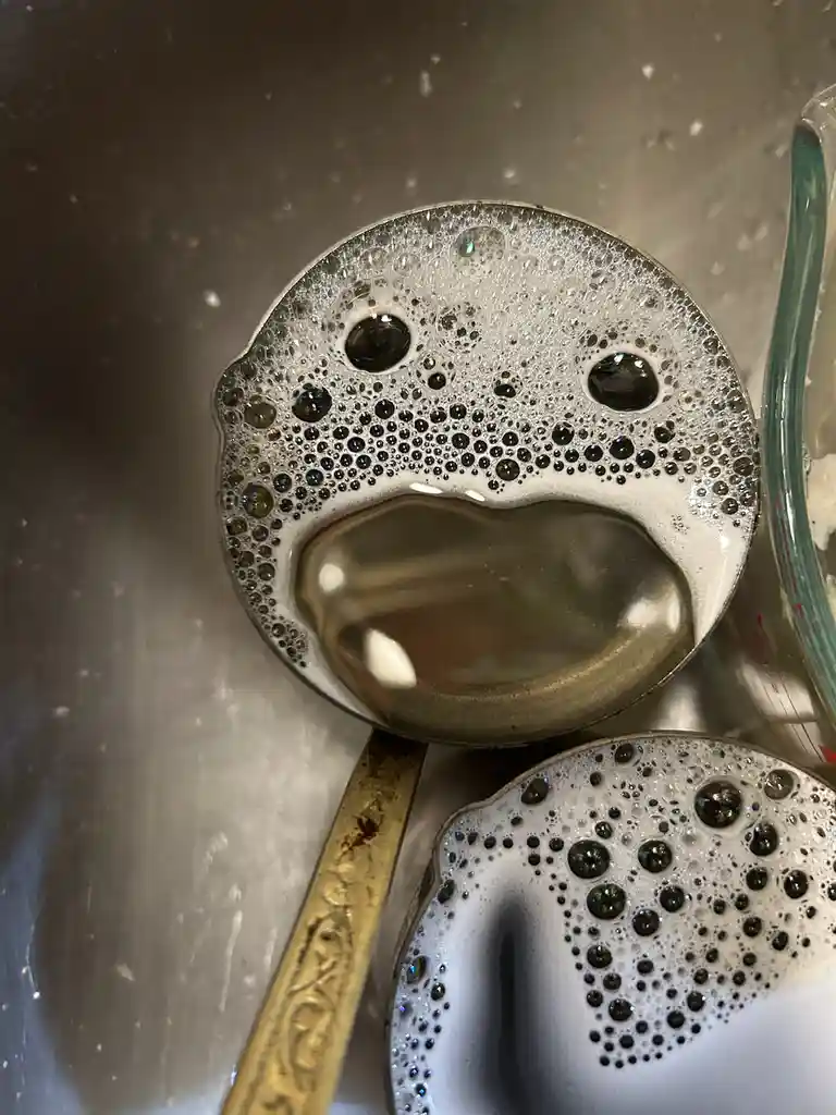 A close-up photo shows two metal spoons in a stainless steel sink, both covered in soapy bubbles that form a sad, crying…