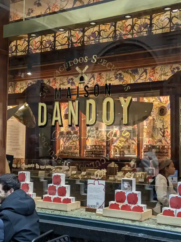 A storefront window of 'MAISON DANDOY' with ornate floral wallpaper and displays of packaged pastries, including boxes…