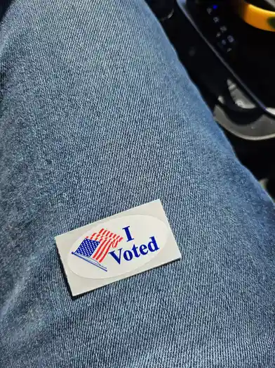 A close-up photo shows a standard American 'I Voted' sticker, featuring a stylized flag, stuck on a person's blue denim…