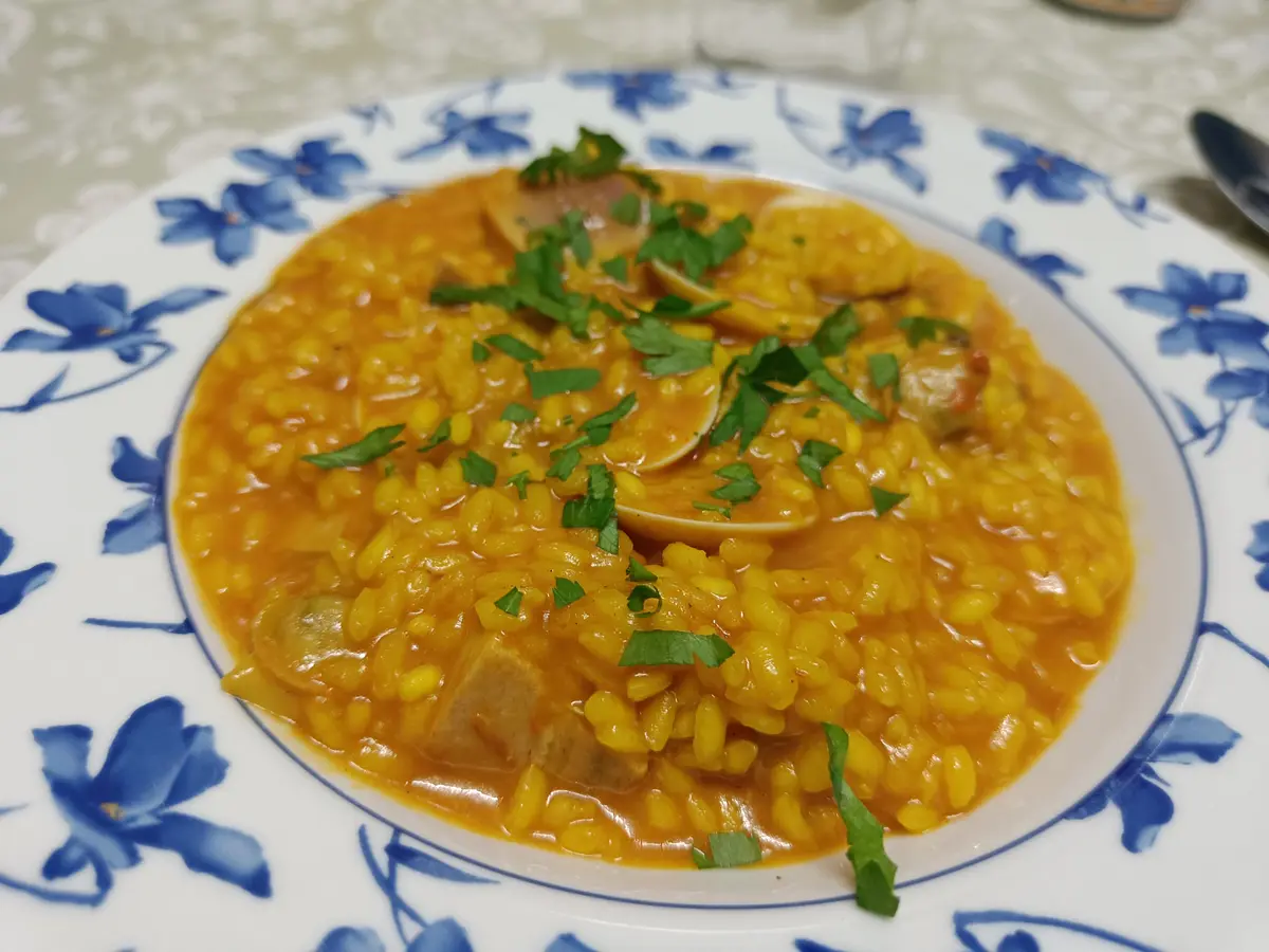A close-up photo shows a white plate with blue floral patterns holding a dish of yellow rice with clams, garnished with…