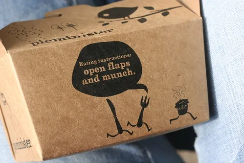 A brown cardboard food box, branded 'pieminter', features a humorous illustration of a fork and knife with legs running away…