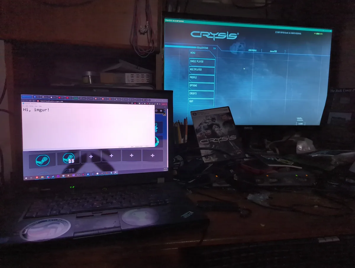 A laptop displays 'Hi, imgur!' in Notepad while a larger monitor behind it shows the Crysis game menu. The setup includes a…
