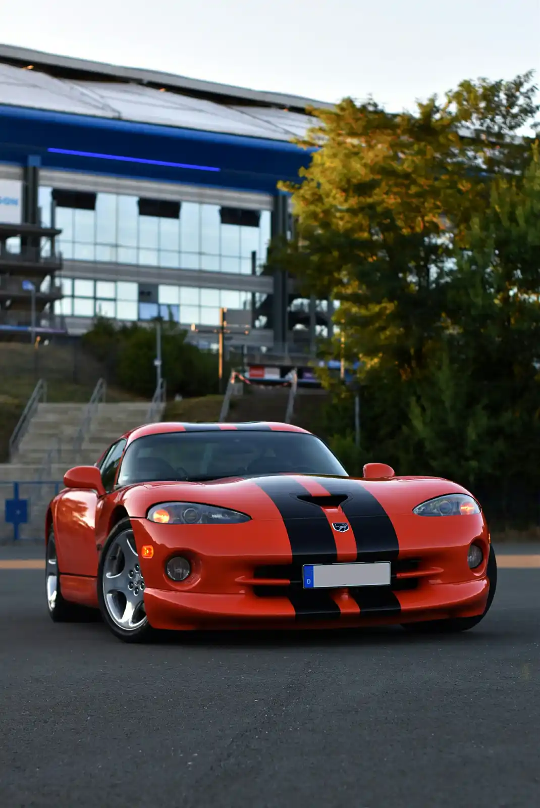 A red Dodge Viper with black racing stripes is parked on asphalt in front of a modern building with glass windows and blue…