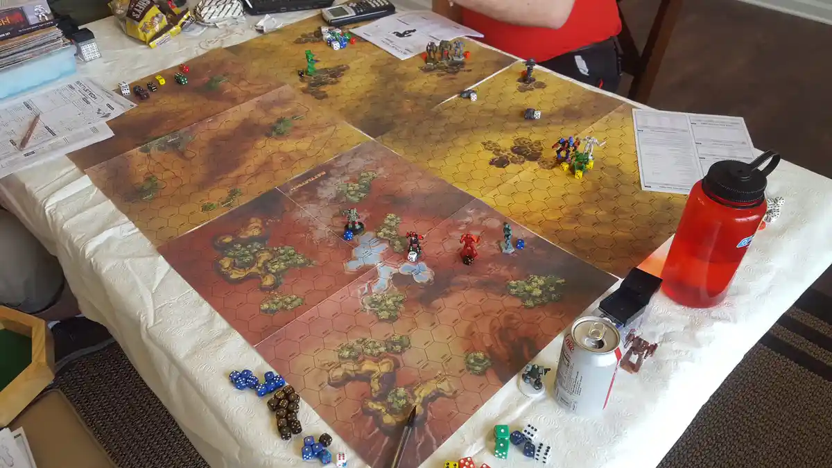 An overhead photo of a BattleTech tabletop wargame in progress, showing multiple hexagonal battle mats with miniature mechs…