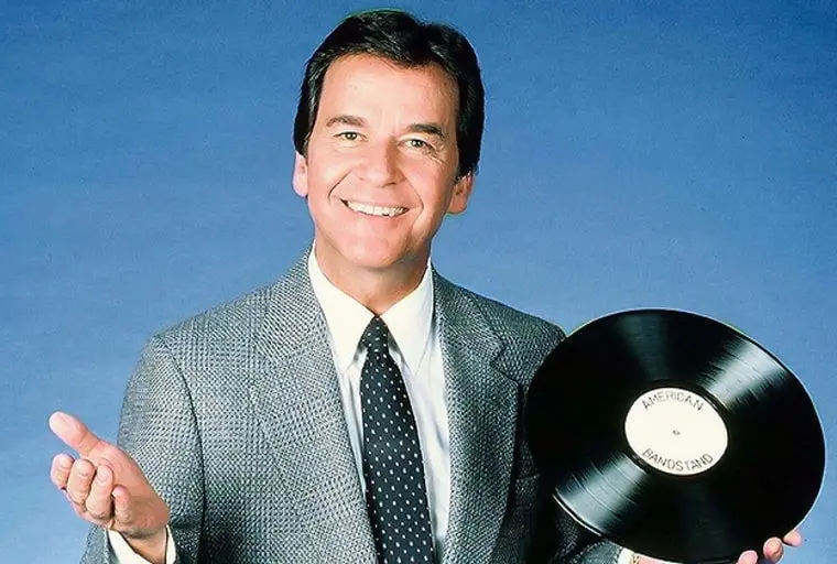 a smiling man in a gray suit and polka-dot tie holds a black vinyl record with 'american bandstand' printed on its label…