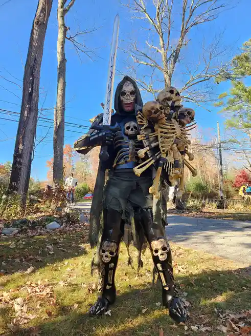 A person in a dark, hooded skeleton costume stands outdoors on grass with fallen leaves, holding a large sword and carrying…