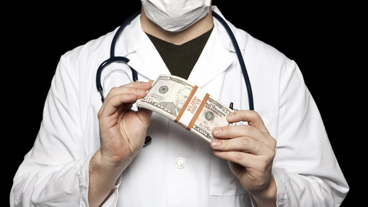 A person wearing a white doctor's coat, a surgical mask, and a stethoscope holds a bundle of US $50 bills tied with a band…