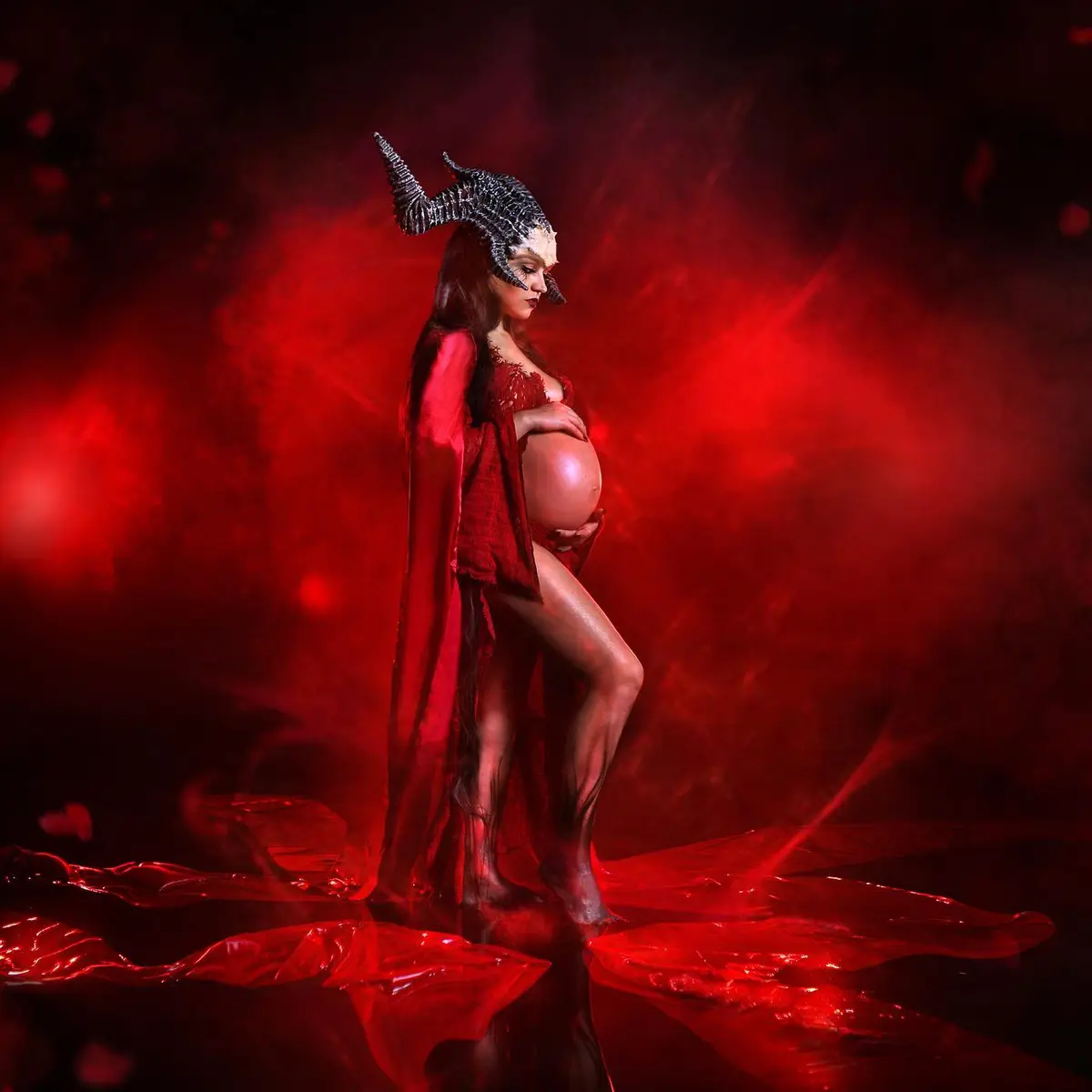 A pregnant woman in a red outfit and black horns stands in a red-lit, smoky environment, holding her belly. The overlaid…