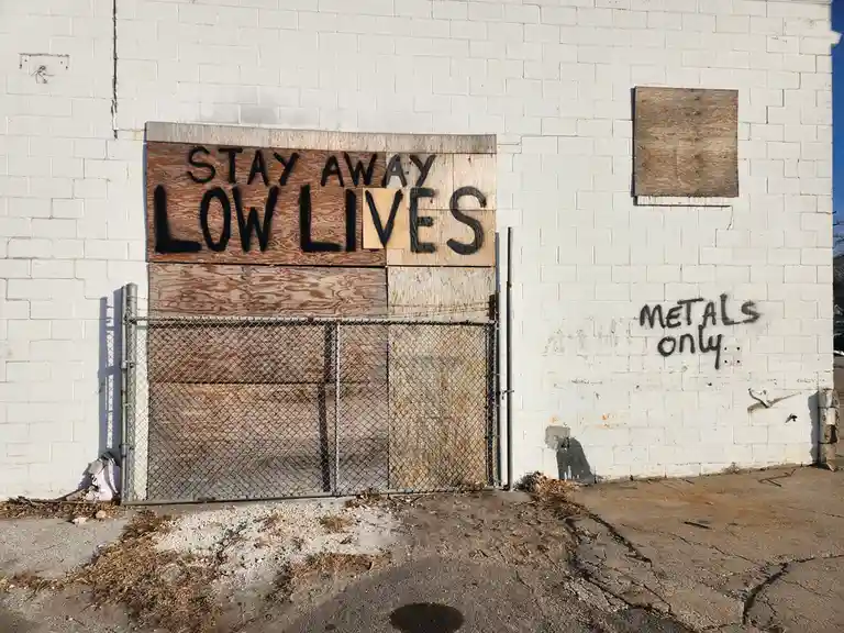 A white brick building has a chain-link gate covered with plywood, with 'STAY AWAY LOWLIVES' spray-painted in black on the…