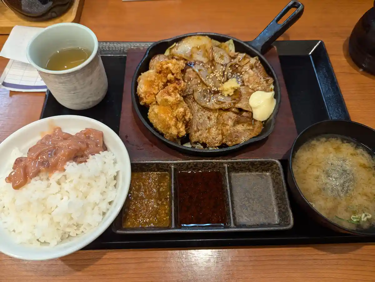 A Japanese meal featuring a cast-iron skillet with grilled meat and fried chicken, a bowl of rice topped with a pink sauce…