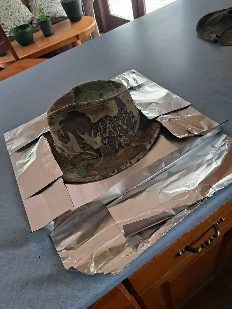 A camouflage-patterned bucket hat sits on a kitchen counter surrounded by crumpled aluminum foil, suggesting an attempt at…