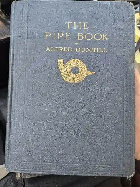 A close-up of a vintage, dark blue cloth-bound book titled 'THE PIPE BOOK' by Alfred Dunhill, featuring a gold-embossed…