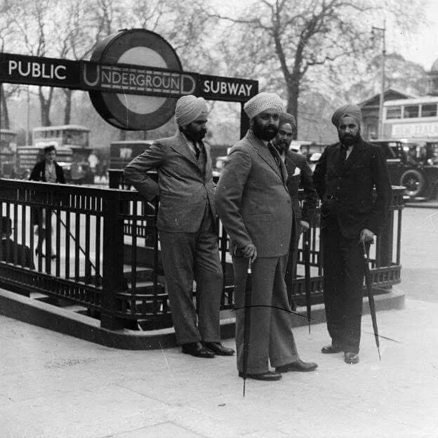 A black-and-white historical photo shows three men in suits and turbans standing confidently near a vintage London…