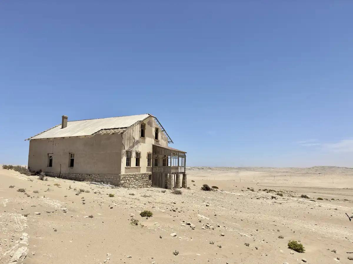 An abandoned, dilapidated two-story house with a corrugated metal roof and stone foundation sits alone in a vast, arid…