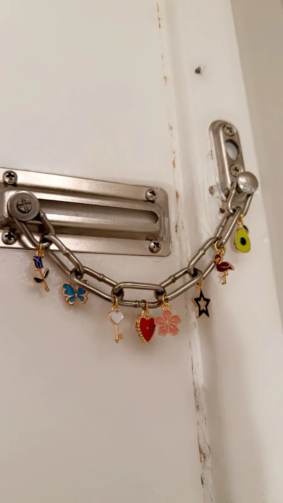 A close-up photo shows a metal door chain lock attached to a white door, decorated with numerous colorful enamel charms…