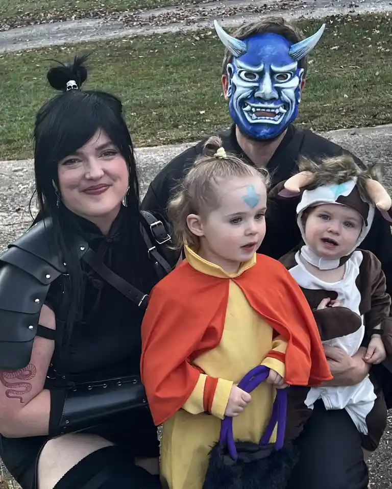 A family of four in costumes poses outdoors: a woman in black armor, a man wearing a blue demon mask, a toddler in an orange…
