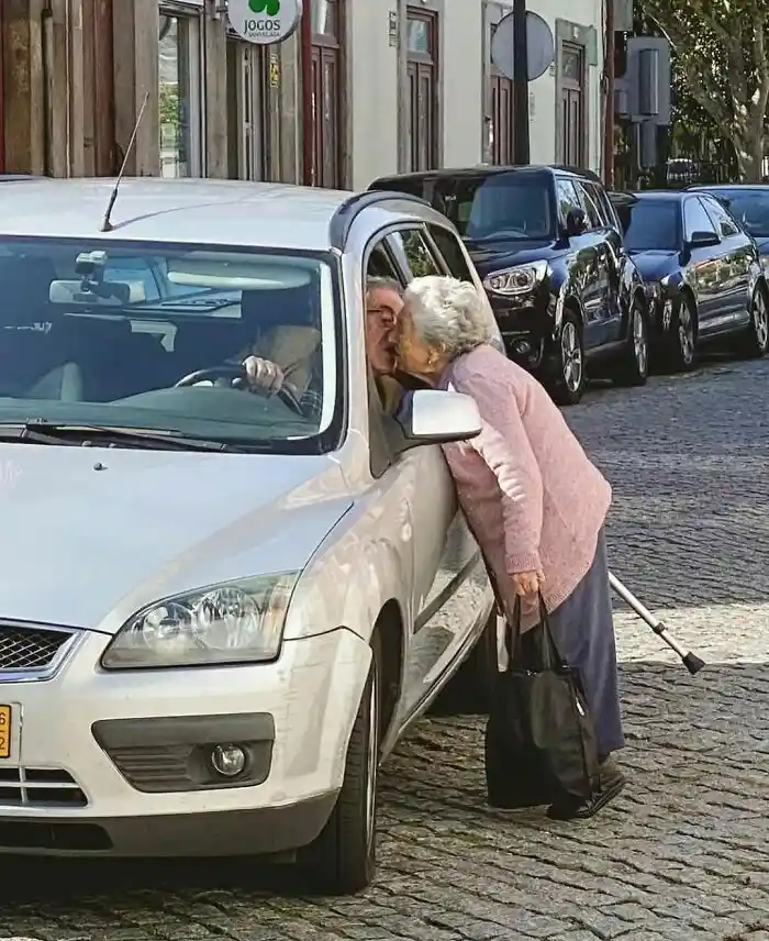An elderly woman with a cane leans into the open window of a silver car, kissing an older man who is driving. The man is…