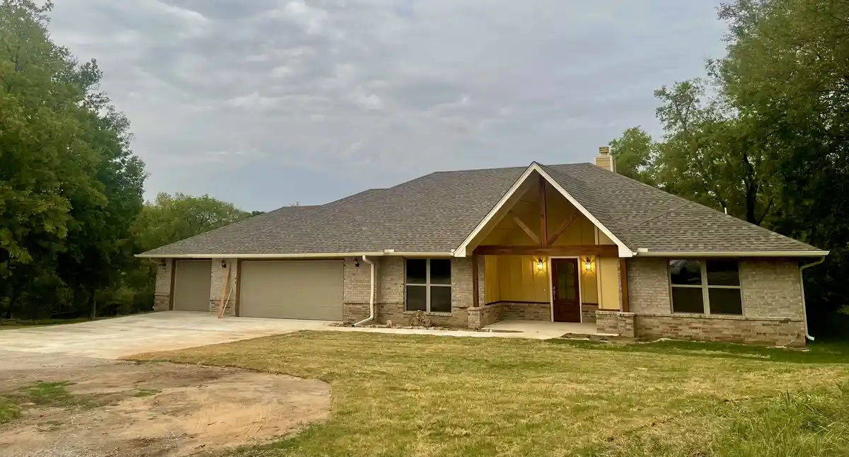A single-story suburban house with brick siding, a gray shingle roof, and two garage doors is shown under an overcast sky…