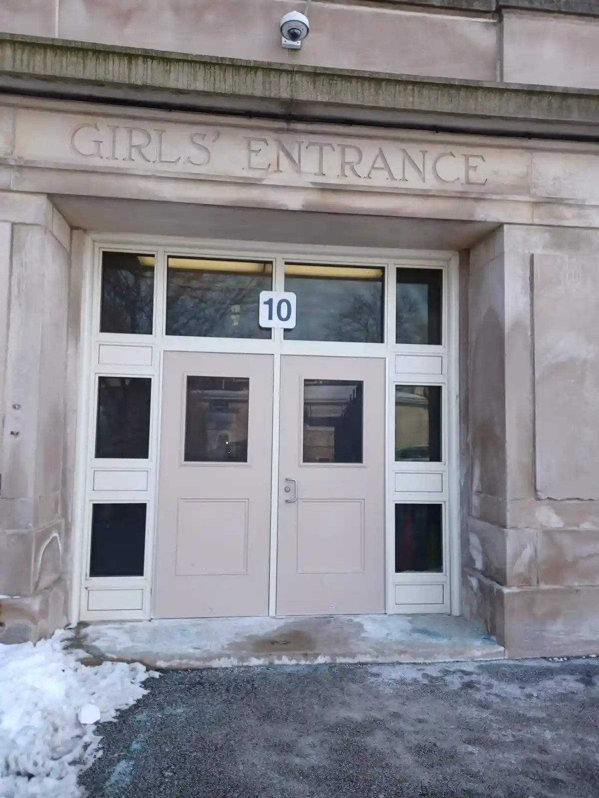 A photo of a building entrance with stone lettering above the doors that reads 'GIRLS' ENTRANCE'. The doors are light pink…