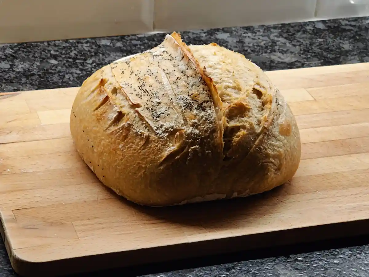 A round, golden-brown loaf of artisan bread with a scored top and poppy seeds sits on a wooden cutting board. The bread has…