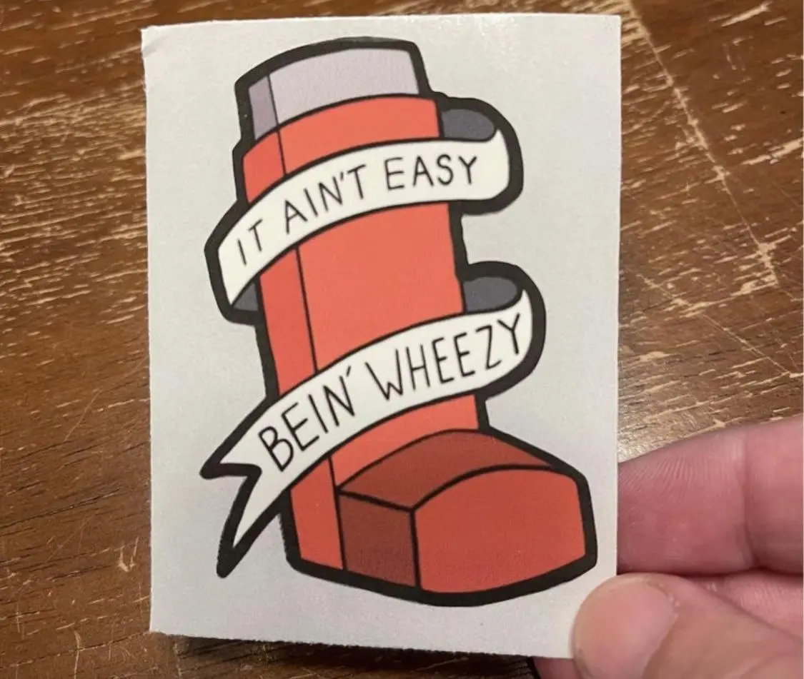 A hand holds a sticker depicting a cartoon asthma inhaler with two white ribbons wrapped around it. The top ribbon reads 'IT…