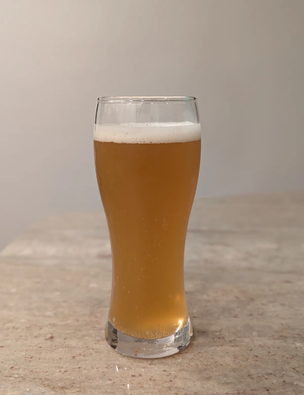 A tall, clear glass filled with golden beer and a white foamy head sits on a speckled countertop against a plain wall. The…