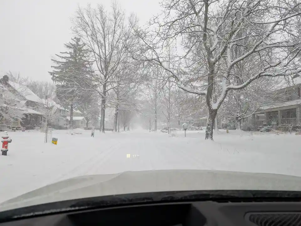A point-of-view shot from inside a car driving down a snow-covered residential street during a blizzard, with bare trees and…