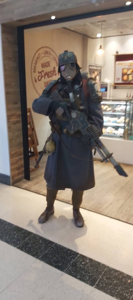 A person in a full-body gas mask and trench coat costume, holding a prop weapon, stands in front of a bakery entrance with a…