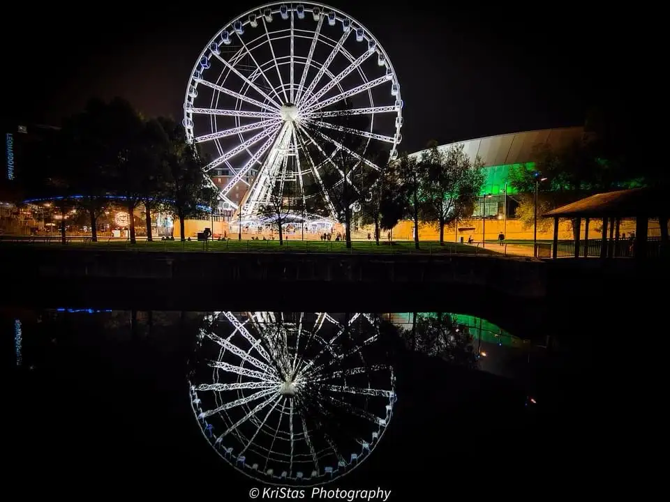 A nighttime photo shows a brightly lit Ferris wheel reflected in dark water, with trees and buildings in the background. The…