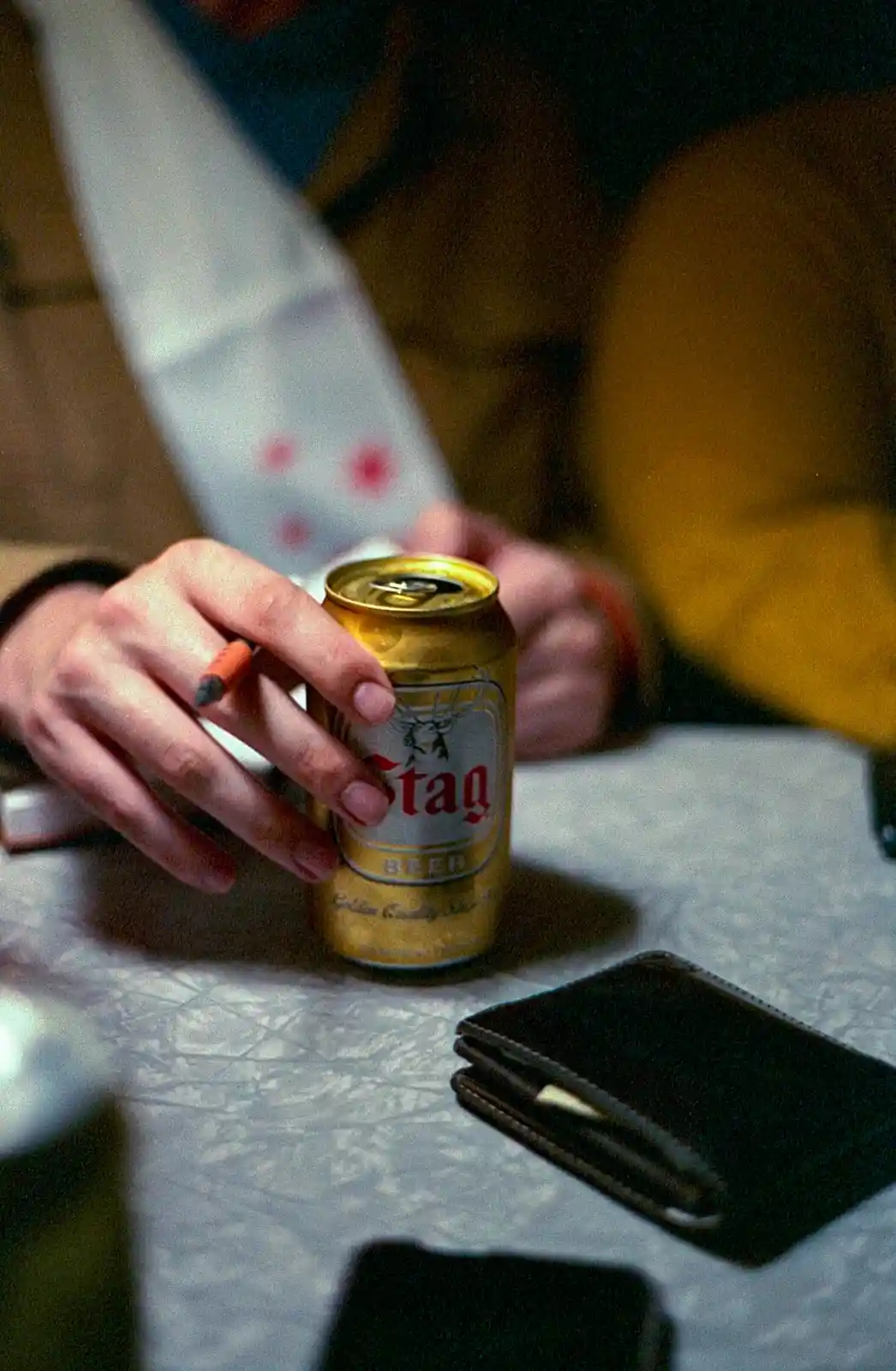 A close-up photo shows a person's hand holding a gold Stag beer can while a lit cigarette rests on their finger. In the…