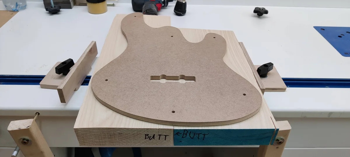 A light-colored wooden guitar body blank is clamped to a workbench, with the word 'BUTT' handwritten on both sides of the…