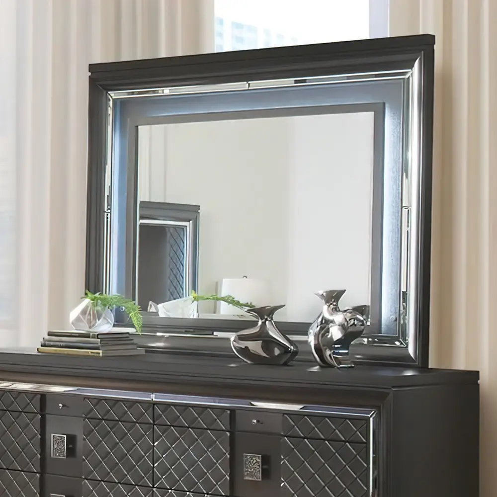 A black-framed mirror with a lighted border sits atop a matching dresser, reflecting its own frame and a second, smaller…