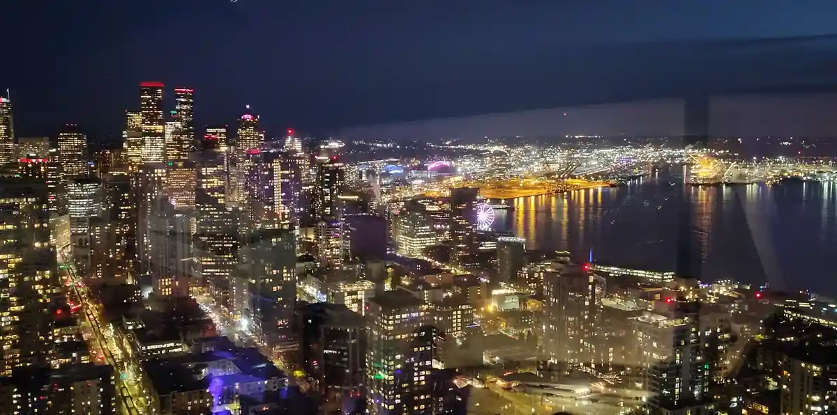 A nighttime cityscape photo of Seattle, showing illuminated skyscrapers, a waterfront, and a Ferris wheel, viewed through a…