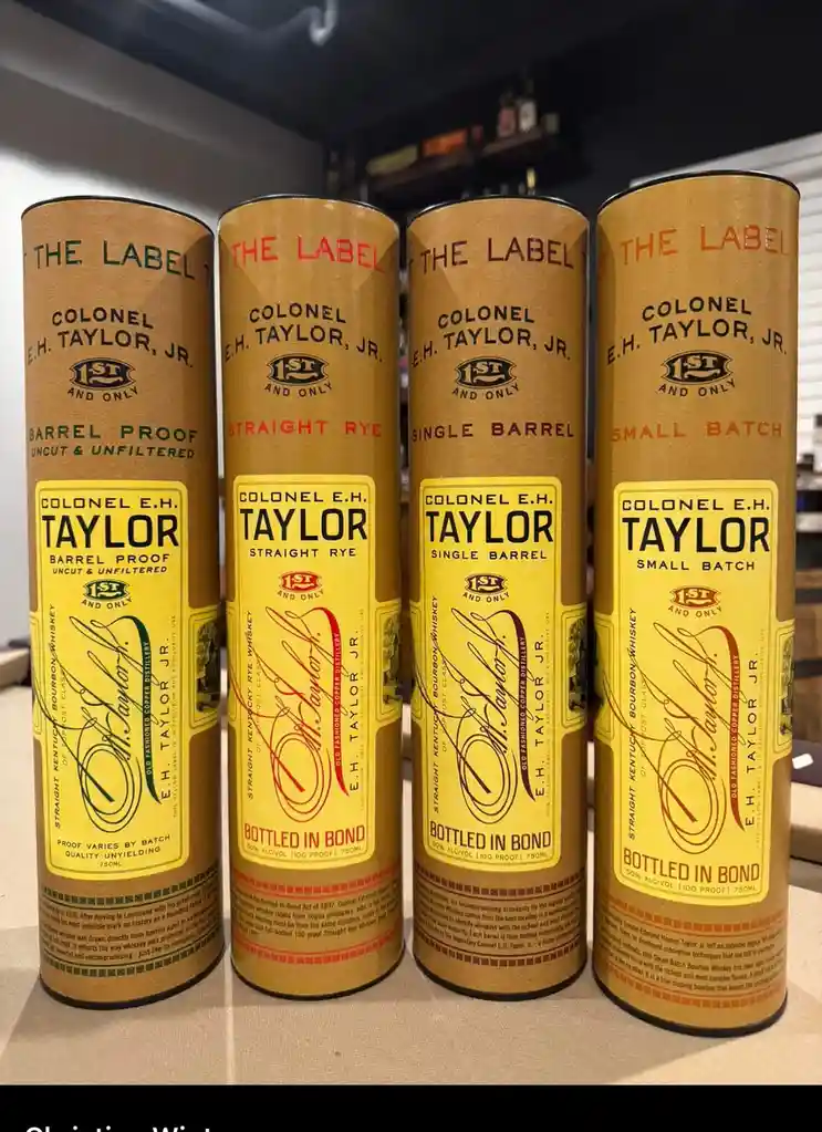 Four identical cylindrical cardboard tubes of Colonel E.H. Taylor, Jr. whiskey are lined up side-by-side, each featuring a…