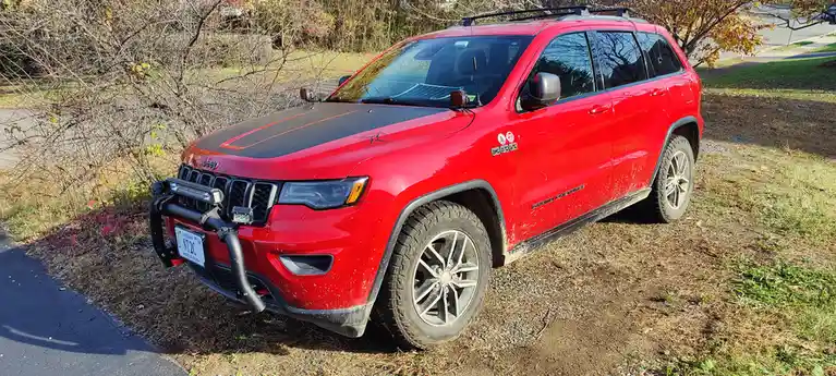 A red Jeep Grand Cherokee is parked on a grassy, gravelly area next to a paved road, with autumn foliage and bare branches…