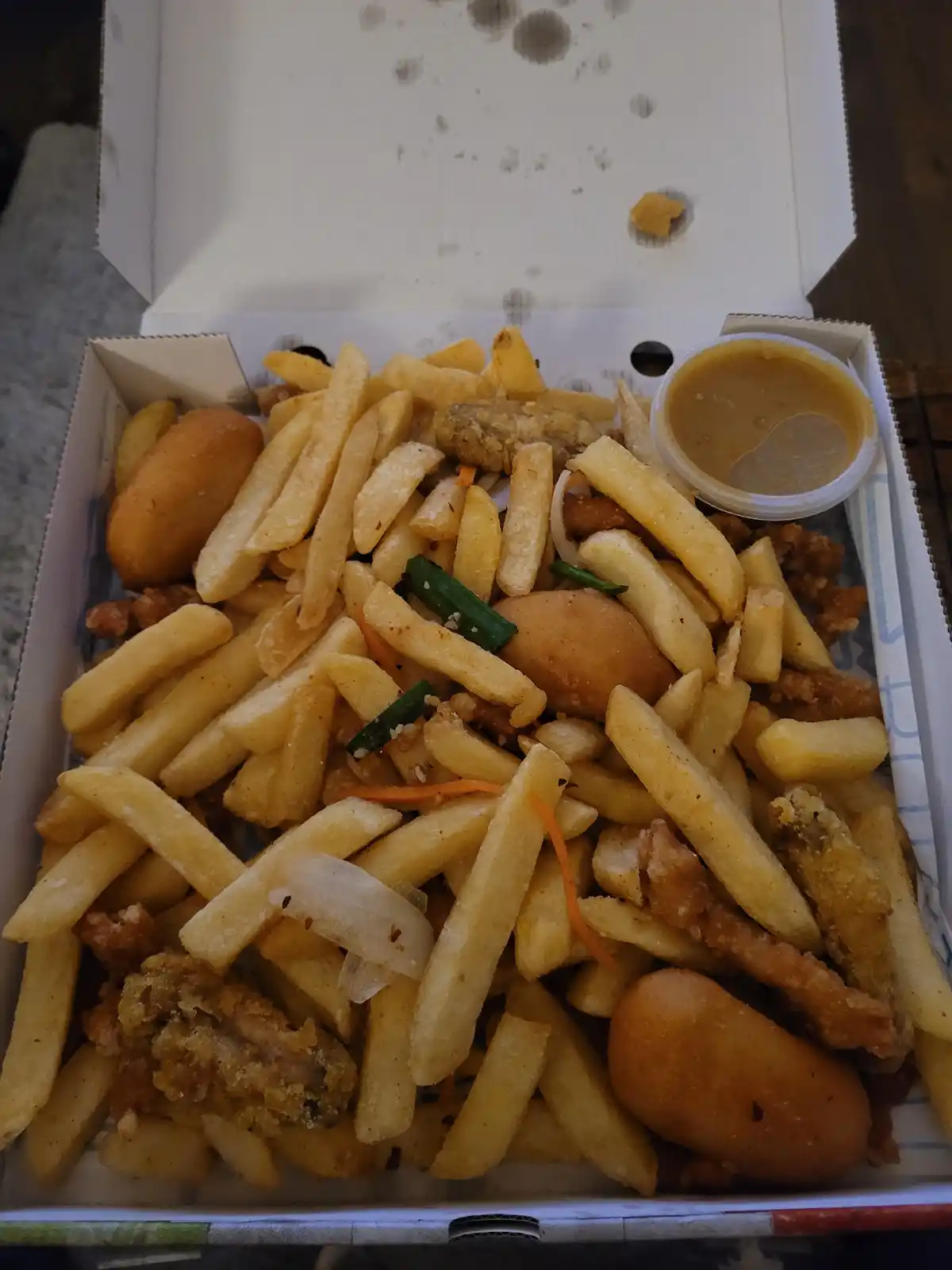 An open cardboard food box is filled to the brim with golden french fries, fried chicken pieces, fried corn dogs, and a…