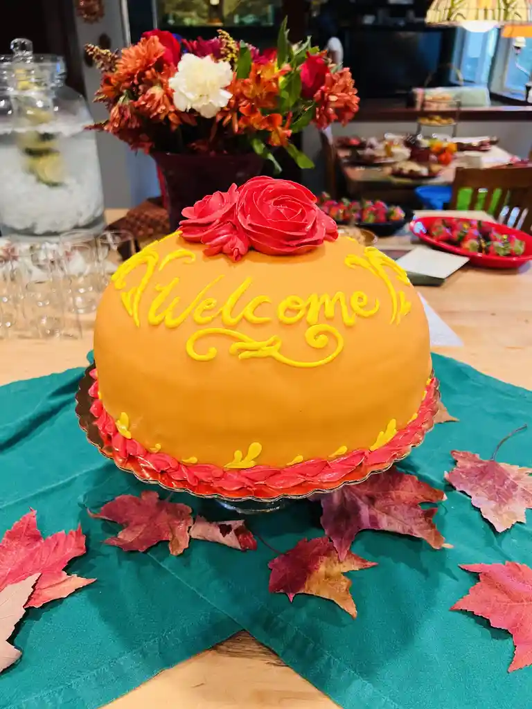 A round, orange-frosted cake with yellow icing spelling 'Welcome' and red frosting roses on top sits on a teal tablecloth…
