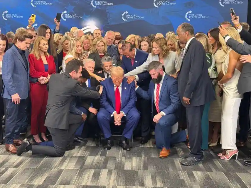 a man in a blue suit and red tie sits in a chair surrounded by a crowd of people, some kneeling and placing hands on his…