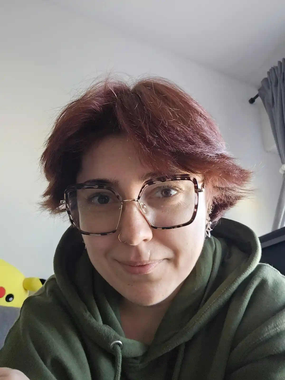 A person with short reddish-brown hair, wearing large tortoiseshell-framed glasses and a green hoodie, looks directly at the…