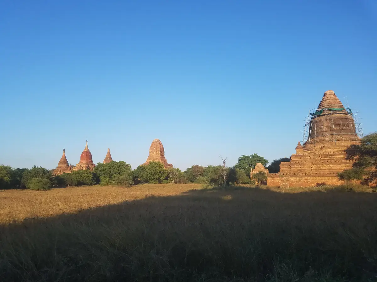 A scenic view of ancient brick temples in Bagan, Myanmar, under a clear blue sky, with one temple on the right covered in…