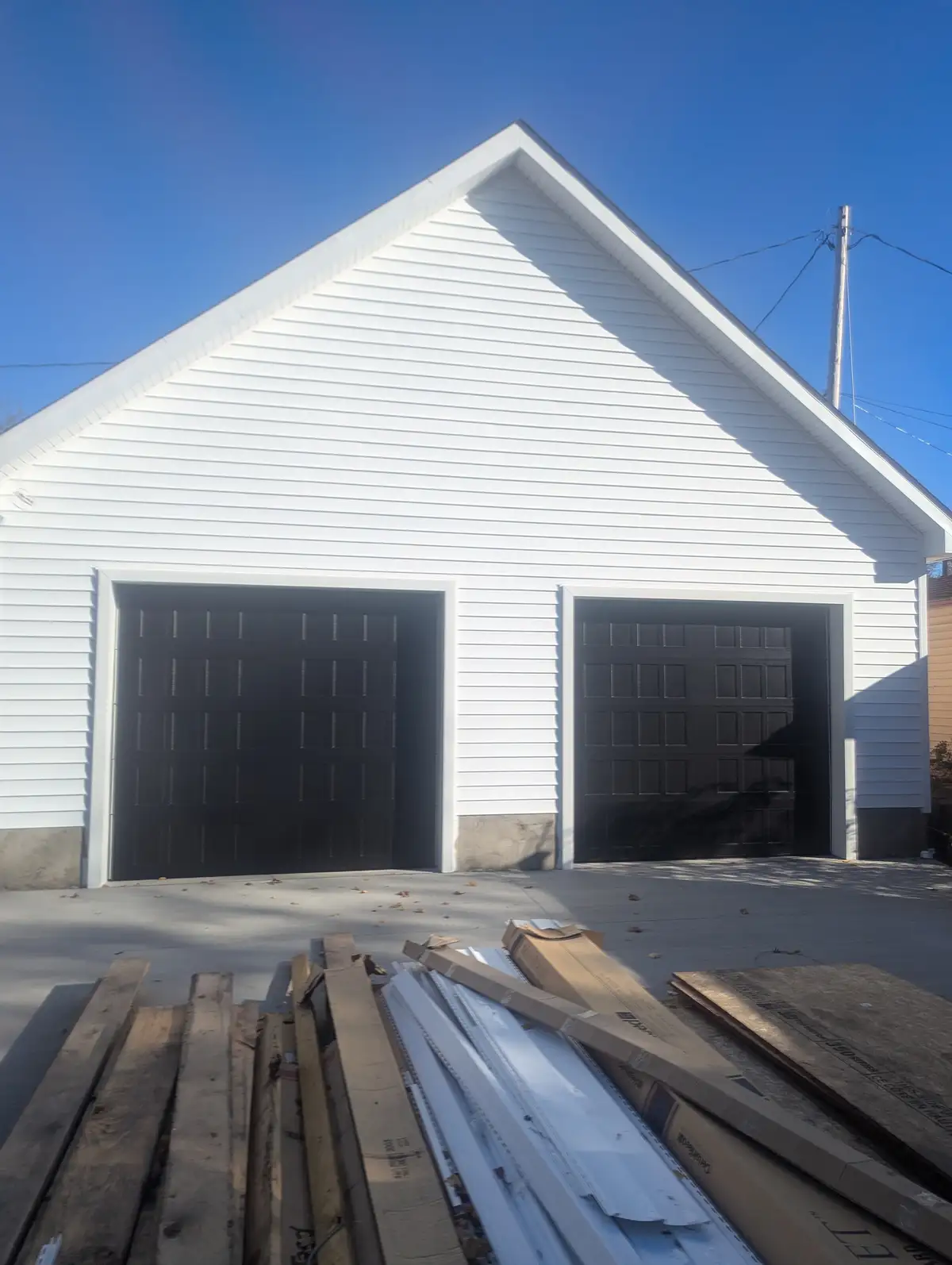A white two-car garage with black doors under a clear blue sky, with construction materials like wood planks and cardboard…