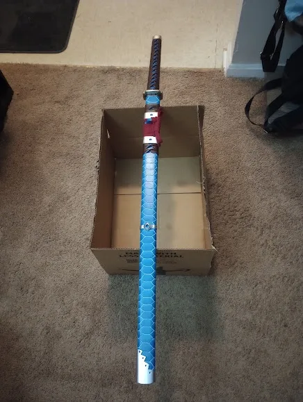 A blue, hexagon-patterned sword with a red and blue hilt rests vertically inside an open cardboard box on a beige carpet…