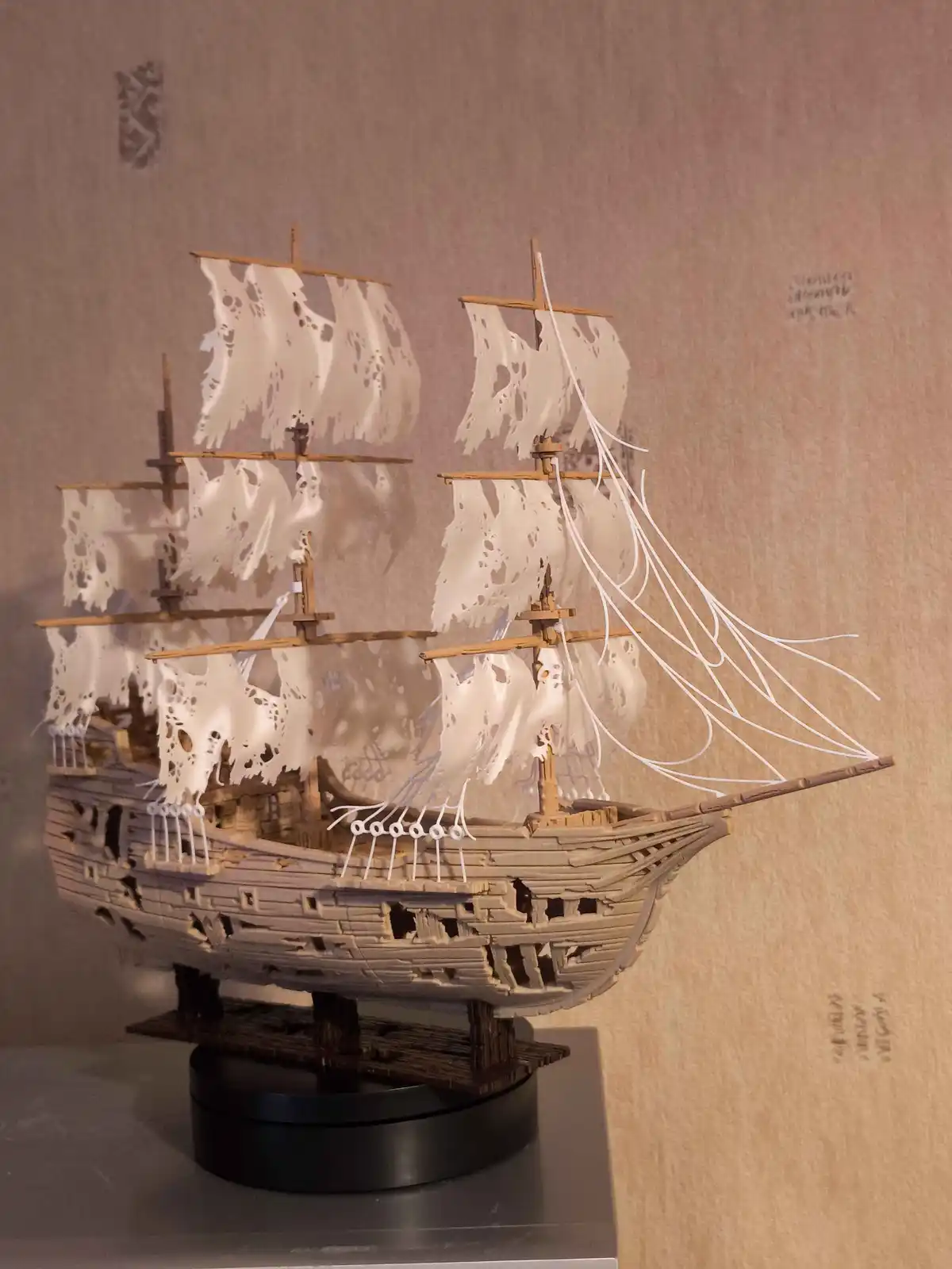 a detailed model of a wooden sailing ship with multiple masts and tattered white sails, displayed on a black stand against a…