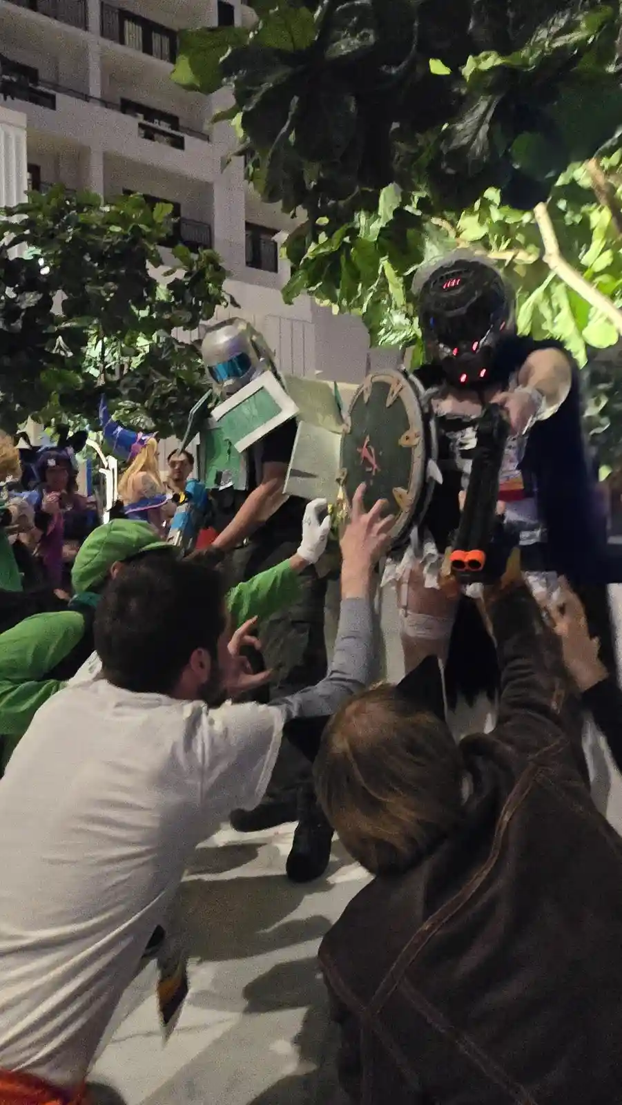 At a nighttime cosplay event, a person in a dark, glowing-eyed costume holds a shield and a toy gun while being surrounded…
