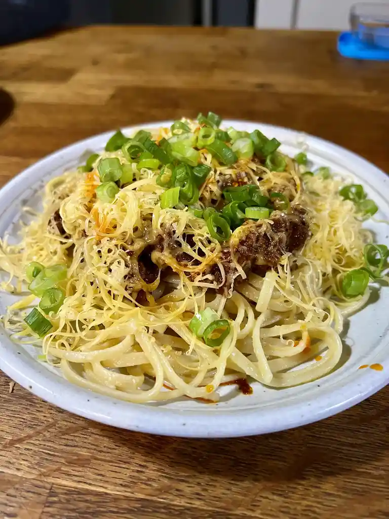 A close-up photo of a plate of pasta with meat sauce, melted cheese, and chopped green onions, sitting on a wooden table…